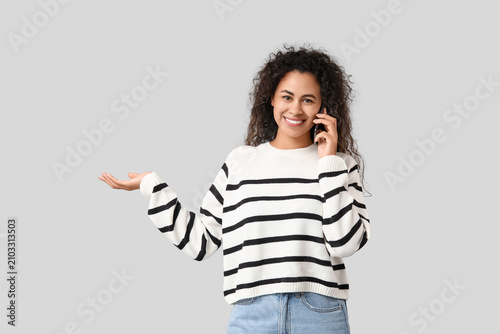 Young African-American woman showing something while talking by phone on grey...