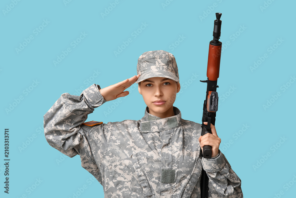 Fototapeta premium Confident young female soldier with assault rifle saluting on blue background