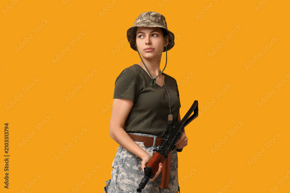 Fototapeta premium Confident young female soldier with assault rifle on yellow background
