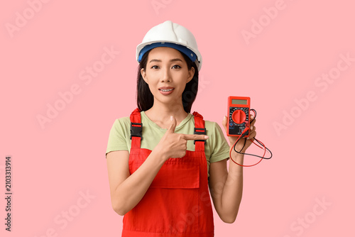 Young female electrician pointing at multimeter on pink background
