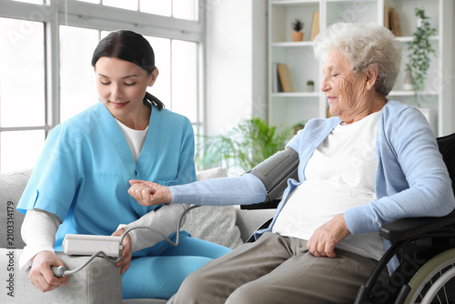 Female nurse measuring blood pressure of senior woman in wheelchair at home