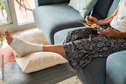 Obraz na płótnie Anonymous woman with bandaged foot solving crossword puzzle while recovering at