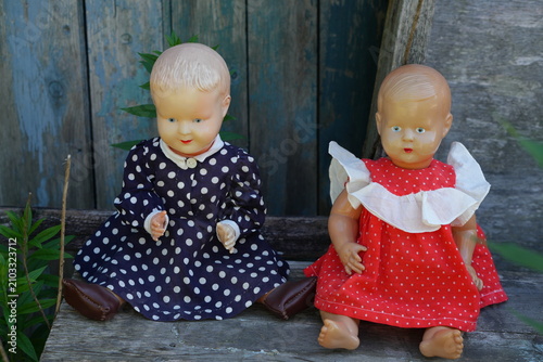 Two vintage plastic baby dolls dressed in polka dot outfits sit side by side on a weathered wooden stool against a rustic blue background