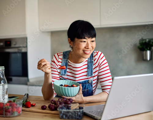 Portrait of a healthy young asian woman enjoying delicious fruit and cereal salad breakfast bowl and using a laptop to work at home