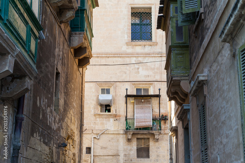 Weathered Valletta buildings with green wooden gallarija balconies and an air conditioner unit line a narrow shaded alley
