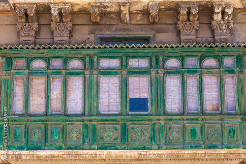 A weathered green wooden enclosed balcony with arched windows and blinds decorates a historic facade in Valletta, Malta