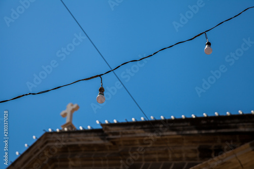 String lights hang above a church rooftop cross against a clear blue sky in Valletta, Malta, during a festival celebration