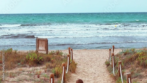 Young girl in white pants and purple top walks along sandy path towards ocean, raising arms in joy as waves crash on shore under cloudy sky