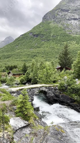 Powerful mountain river rushing beneath a historic stone bridge, surrounded by majestic mountains and pristine Norwegian wilderness. Crystal-clear water, rugged rocks, and breathtaking Scandinavian sc
