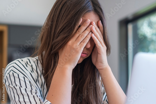 Fotografie Stressed woman covering her face with hands while working on a laptop at home, showing frustration, fatigue and overwhelm, concept of remote work stress, burnout and mental health strain