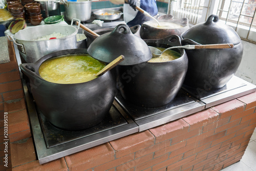 In a local kitchen, large black pots simmer on the stove filled with ingredients soup for a lunchtime meal. Cooking staff prepare food.