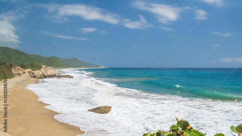 Obraz premium Horizontal image. Tropical beach with waves breaking on sand and large smooth rocks under a blue sky