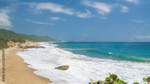 Horizontal image. Tropical beach with waves breaking on sand and large smooth rocks under a blue sky