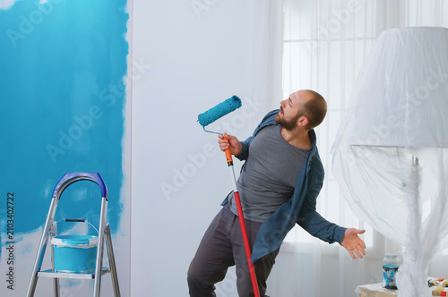 Canvas-taulu Happy repairman sings into a roller brush during home renovation, using blue paint while turning apartment decorating into a playful DIY moment