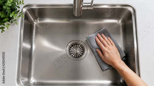 A person is cleaning a shiny stainless steel kitchen sink with a gray cloth next to a green leafy plant.