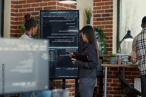 Engineer explains wall screen code errors as engineers use a tablet and wall display to review bugs, cloud computing and team software development in an IT office.