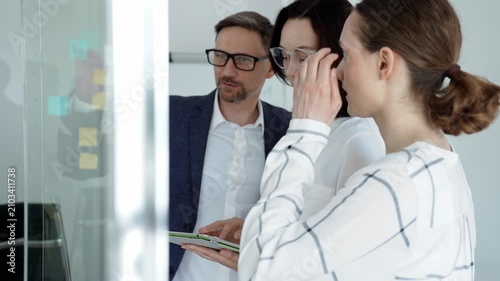 Business team collaborating on a project, analyzing data on a tablet, planning strategy with sticky notes on a glass whiteboard in a modern office meeting