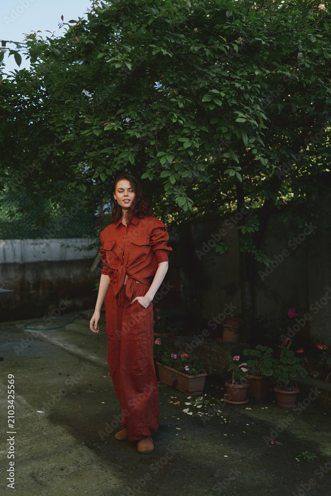 Obraz premium Vibrant lifestyle moment with a woman in a red dress outdoors, relaxed pose, soft smile, sunlit scene, leafy background, natural colors, casual elegance