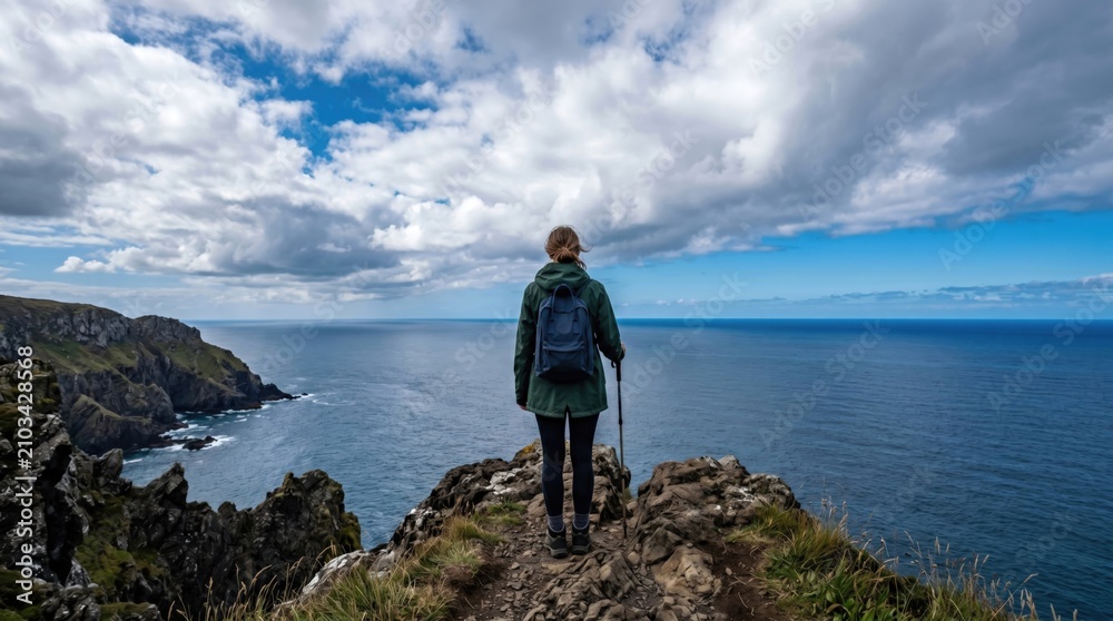 Fototapeta premium Young woman hiker standing on a rocky cliff edge overlooking the vast ocean, representing freedom, adventure, travel, solitude, and personal exploration goals