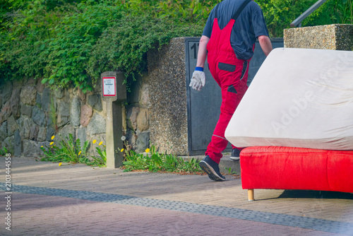 Принти на полотні Worker in red overalls carrying a mattress on a sidewalk near a stone wall and g