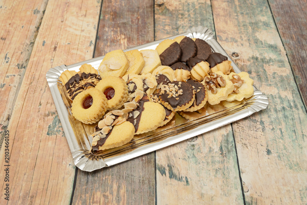 Fototapeta premium Variety of shortbread biscuits including chocolate-dipped, nut-topped, and jam-filled cookies arranged on a silver tray on wood.