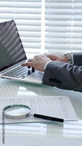 Businessman's hands are actively typing on a laptop keyboard, researching data and analyzing financial documents laid out on a clear desk with a magnifying glass in an office setting