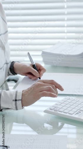 Financial professional performing calculations and writing data, preparing tax forms, managing budget, or completing administrative tasks on a bright office desk with stacks of documents