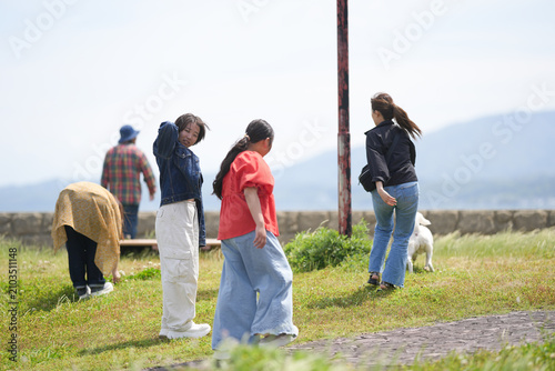 Young women stroll through grassy field with dog nearby. One covers face with hand, appearing thoughtful or tired. Bright sky and distant hills frame casual, relaxed outdoor moment