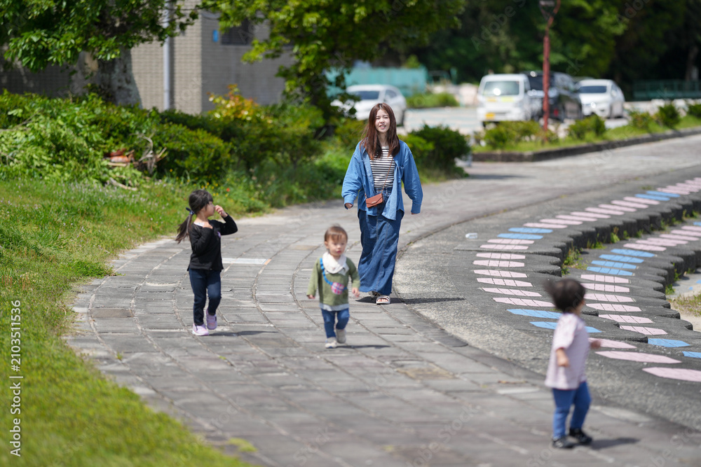 Fototapeta premium Young children play on a curved stone path. Woman walks nearby, smiling gently. Green grass and trees frame the scene. Bright sunlight enhances the peaceful mood. Perfect for family, education