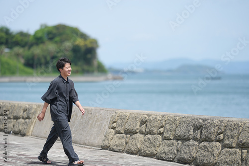 Man walks calmly along seaside stone path. He smiles, enjoying quiet coastal air. Background shows gentle waves and distant green hills. Ideal for travel, wellness, or calm lifestyle themes