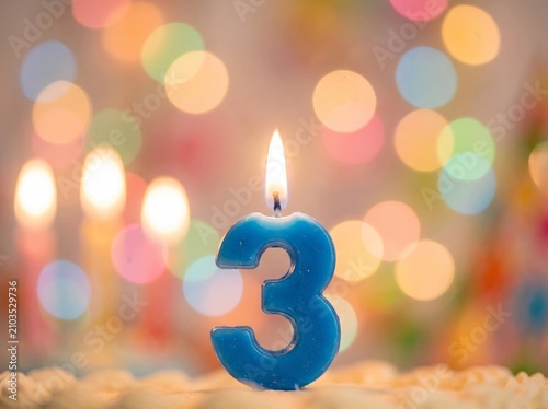 Blue number three birthday candle burning on a cake with beautiful colorful bokeh background for a third anniversary or child's party