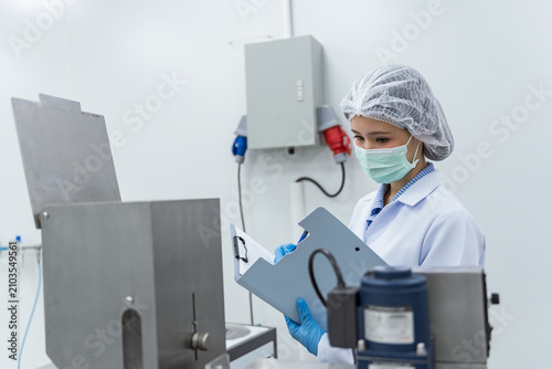 Woman in lab coat, mask, and hairnet examining device. Quality control inspector and factory workers operating automated packaging machinery in a sterile pharmaceutical manufacturing facility.