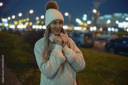 A woman stands outside on a city street during the evening, wearing a warm jacket and a hat with a pompom. She smiles while surrounded by twinkling street lights and distant cars