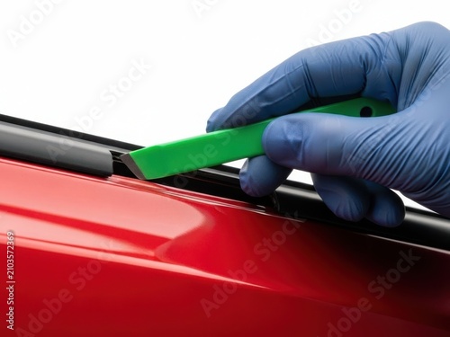 Automotive trim removal tool used on red car body, professional mechanic hand in blue glove prying plastic molding, car detailing and interior disassembly process for vehicle repair