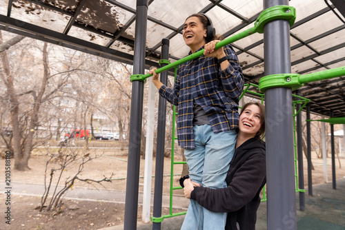 A young woman helps her male friend perform pull-ups at an outdoor gym. Fun and supportive student life.