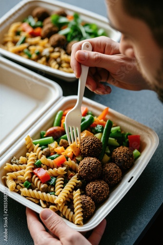Man eats healthy lunch from paper box. Male holds plastic fork for dinner of pasta with falafel plus fresh vegetables. Person sits at table for fast food takeaway meal. Worker enjoys nutritious