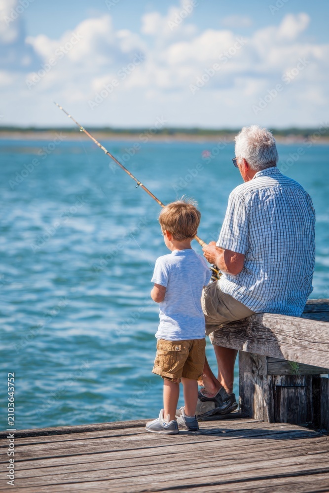 Fototapeta premium Man teaches a young boy to fish from a wooden pier during a sunny day by the water in a coastal area with blue skies and calm waves