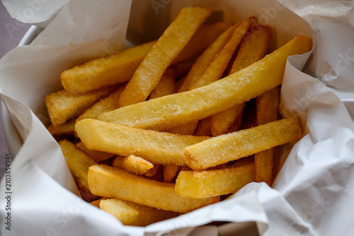 Close up of crispy golden french fries salted and served in a bowl lined with white paper. Delicious fast food snack concept. High angle view with selective focus on food.