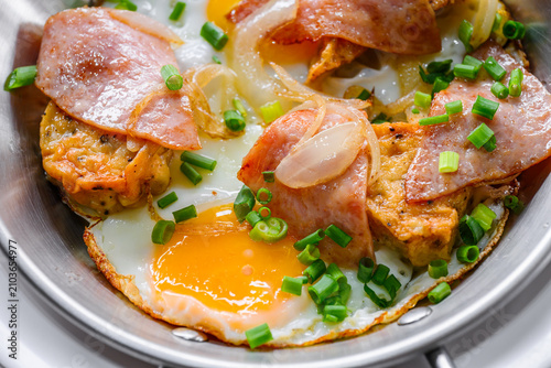 Close up of pan fried eggs topped with ham, sauteed onions, and chopped green onions served in a stainless steel skillet. Delicious Asian breakfast concept. Food close up.