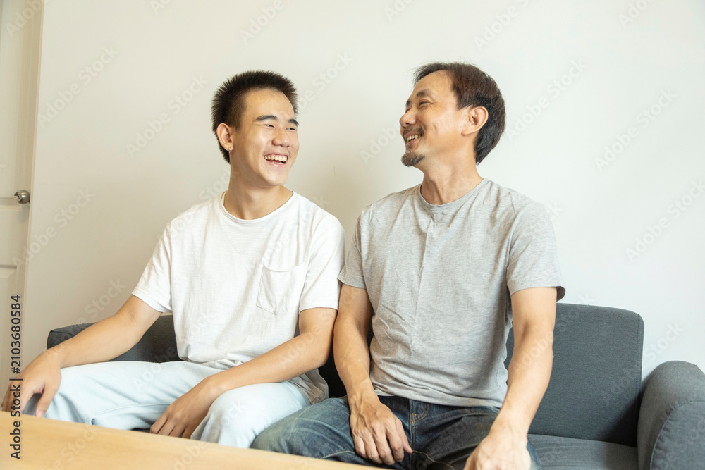 Obraz premium Happy Asian father and son sitting on sofa at home. Cheerful father with teenage son sitting on couch in the living room of apartment. Portrait of father and son spending leisure time at home