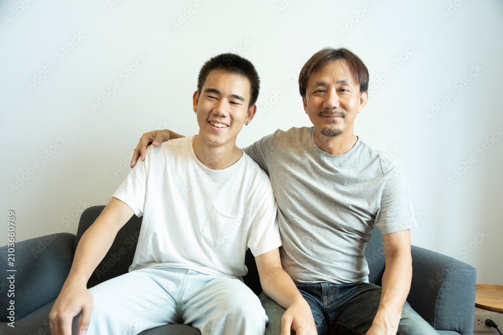 Obraz premium Happy Asian father and son sitting on sofa at home. Cheerful father with teenage son sitting on couch in the living room of apartment. Portrait of father and son spending leisure time at home