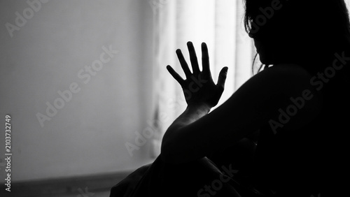 Silhouette of a woman sitting in a dark room, raising one hand in a gesture of defense or hesitation against a bright window backdrop