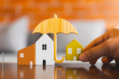 Hand protecting wooden house models under a wooden umbrella. Concept of real estate insurance, home safety, property loan security, and asset protection plan.