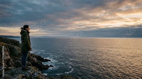 Woman standing on a rocky cliff overlooking the ocean at sunset