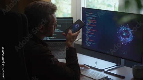 Rear view of coder sitting at desk, using AI voice assistant on a smartphone, giving tasks to artificial intelligence agent on computer, which generates program code. Man interacts with AI chatbot app
