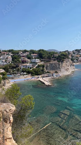 panoramic view of the turquoise waters of Advocat cove in Benissa featuring its iconic breakwater and beach bar along the rocky Mediterranean coastline under a clear blue summer sky