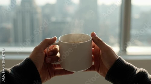 Person holding warm white coffee cup indoors. Silhouette enjoying hot beverage by bright window view.