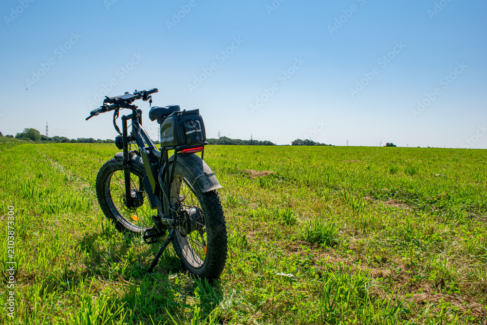 Fototapeta premium Rear view of an electric mountain bike with thick fat tires parked in a vast green meadow under a clear blue sky 