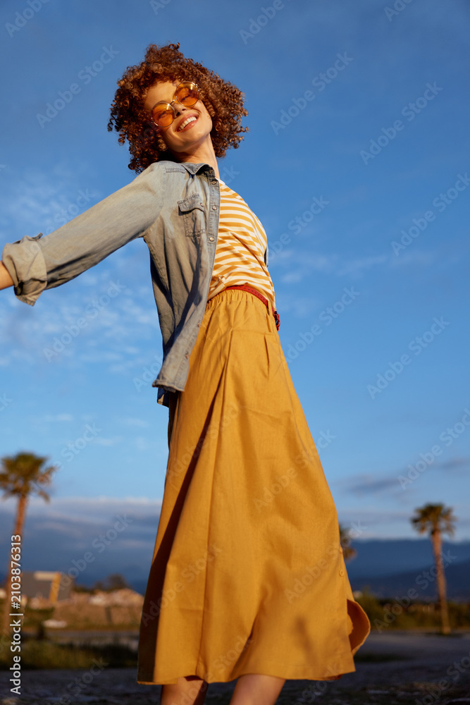 Fototapeta premium Smiling woman in denim jacket and striped top twirls outdoors under bright sky, flowing orange skirt. Lifestyle, outdoor photography, carefree mood and expressive movement.