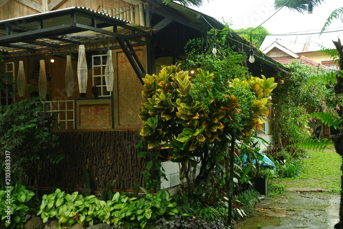 Rustic wooden coffee shop exterior featuring vibrant variegated croton plants and hanging woven bamboo lanterns under a covered porch roof during a bright daylight afternoon in a lush tropical garden 